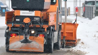 Orangefarbener Schneeräum-LKW mit Räumschild fährt auf schneebedeckter Straße und streu
